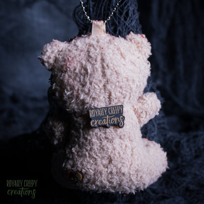 A plush teddy bear ornament or keychain in fuzzy fabric, hanging from a silver chain against a dark textured background. A small badge on the back reads 'ROYALLY CREEPY creations'.