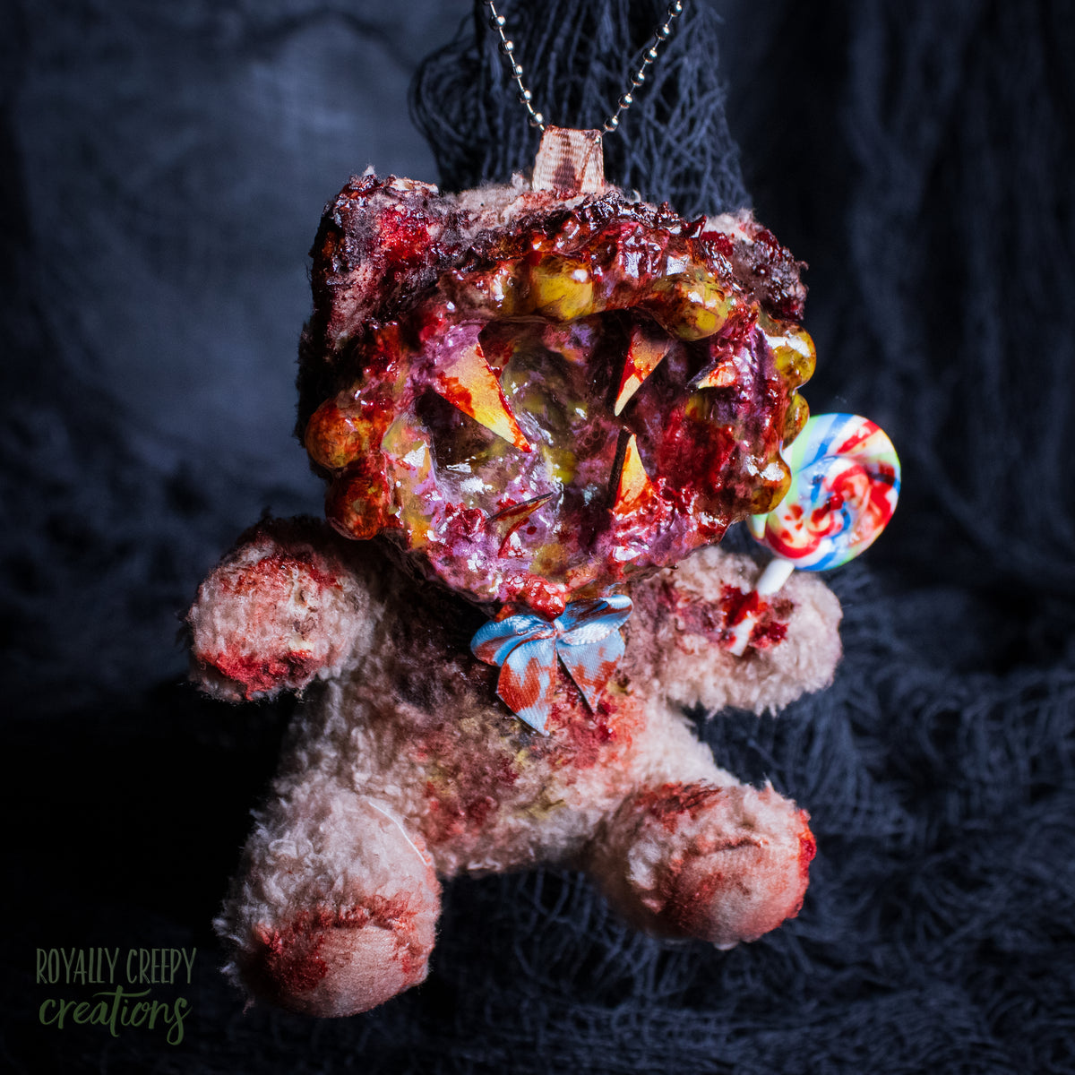 A horror-themed plush bear keychain with a bloody, gory mouth design, fuzzy body, and lollipop accessory, shown against a black netted backdrop.
