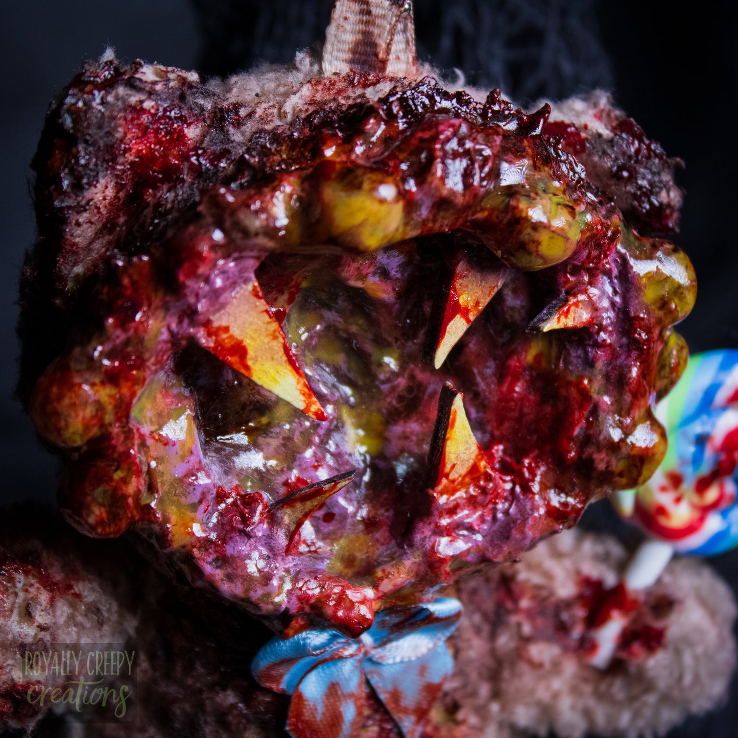 A close-up of a horror-themed plush bear keychain with a bloody, gory mouth design, fuzzy body, and lollipop accessory, shown against a black netted backdrop.
