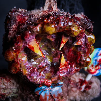 A close-up of a horror-themed plush bear keychain with a bloody, gory mouth design, fuzzy body, and lollipop accessory, shown against a black netted backdrop.