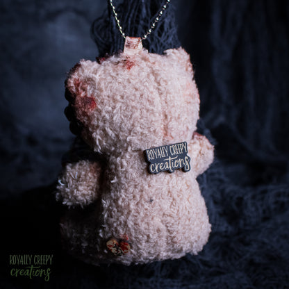A small fuzzy teddy bear shown hanging from a chain against a dark textured background. The bear has distressed details, blood-like red stains, and a small black tag reading 'ROYALLY CREEPY creations'.