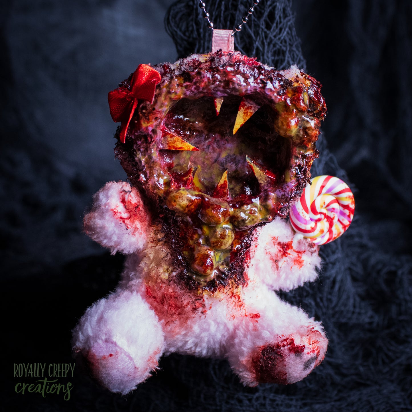 A horror-themed plush bear keychain with a bloody, gory mouth design, fuzzy body, and lollipop accessory, shown against a black netted backdrop.
