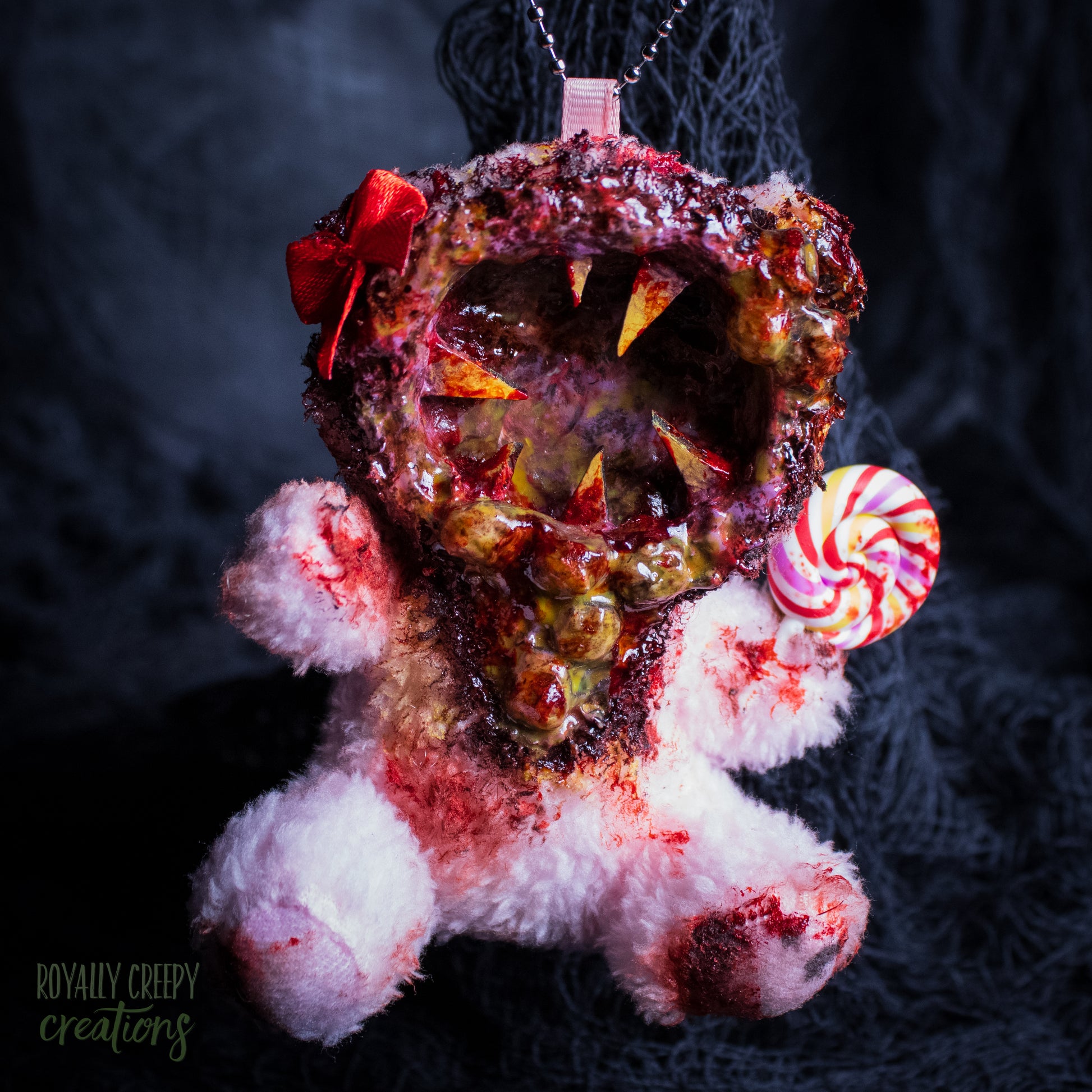 A horror-themed plush bear keychain with a bloody, gory mouth design, fuzzy body, and lollipop accessory, shown against a black netted backdrop.