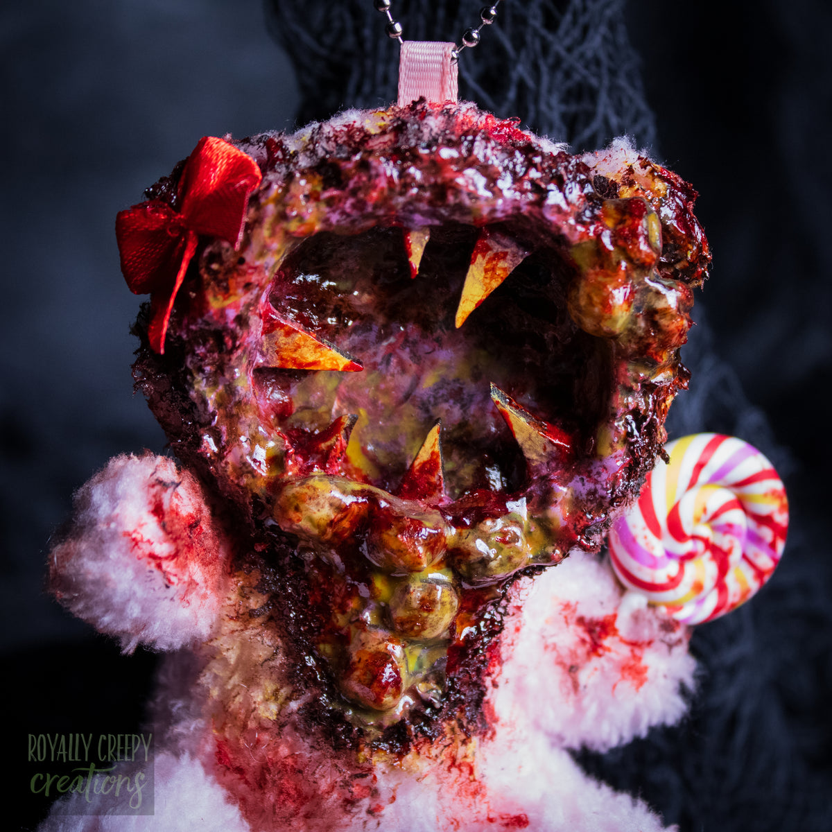 A close-up of a horror-themed plush bear keychain with a bloody, gory mouth design, fuzzy body, and lollipop accessory, shown against a black netted backdrop.