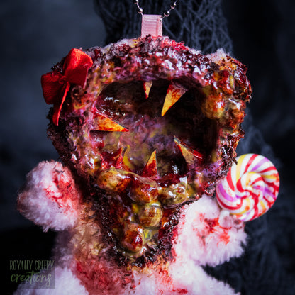 A close-up of a horror-themed plush bear keychain with a bloody, gory mouth design, fuzzy body, and lollipop accessory, shown against a black netted backdrop.