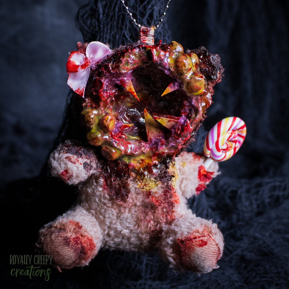 A horror-themed plush bear keychain with a bloody, gory mouth design, fuzzy body, and lollipop accessory, shown against a black netted backdrop.