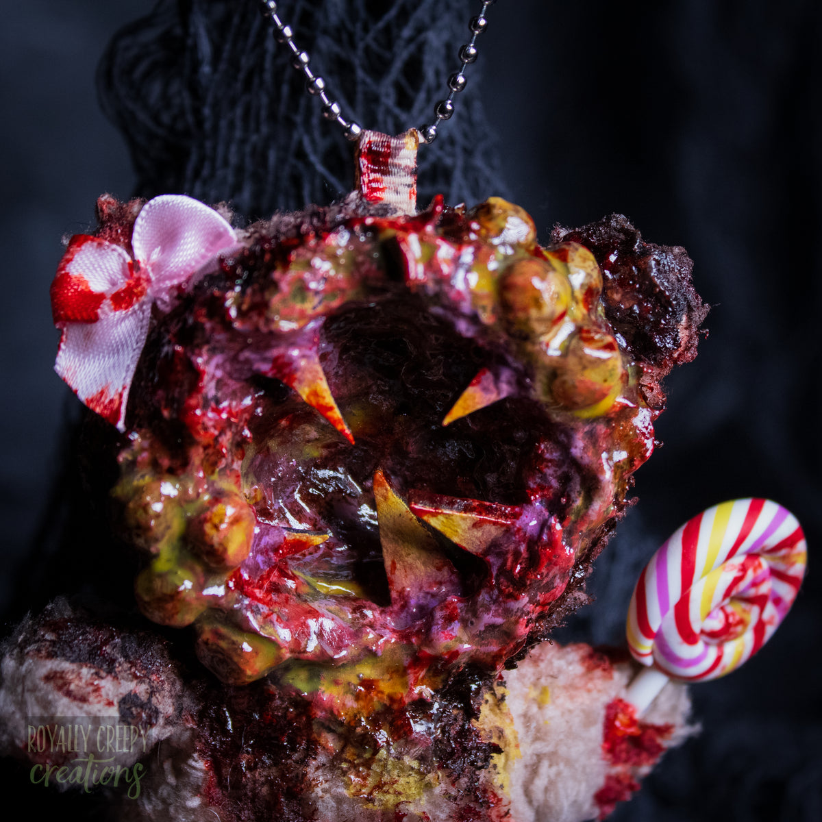 A close-up of a horror-themed plush bear keychain with a bloody, gory mouth design, fuzzy body, and lollipop accessory, shown against a black netted backdrop.