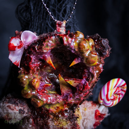 A close-up of a horror-themed plush bear keychain with a bloody, gory mouth design, fuzzy body, and lollipop accessory, shown against a black netted backdrop.