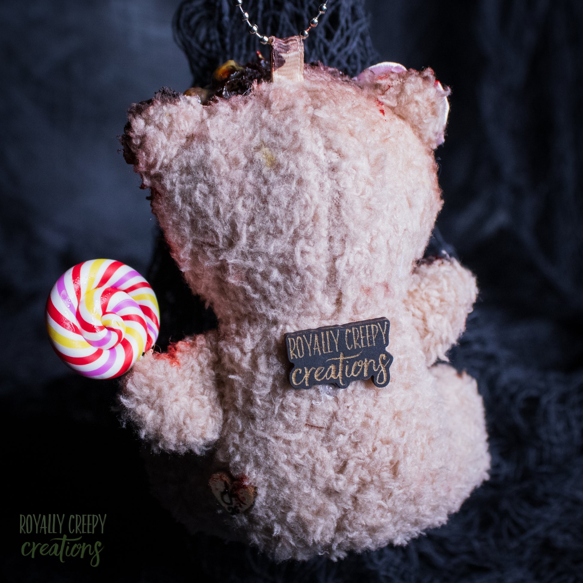 A horror-themed plush teddy bear ornament in fuzzy fabric, holding a colorful spiral lollipop and featuring a 'Royally Creepy Creations' brand tag on its back, set against a dark textured background.