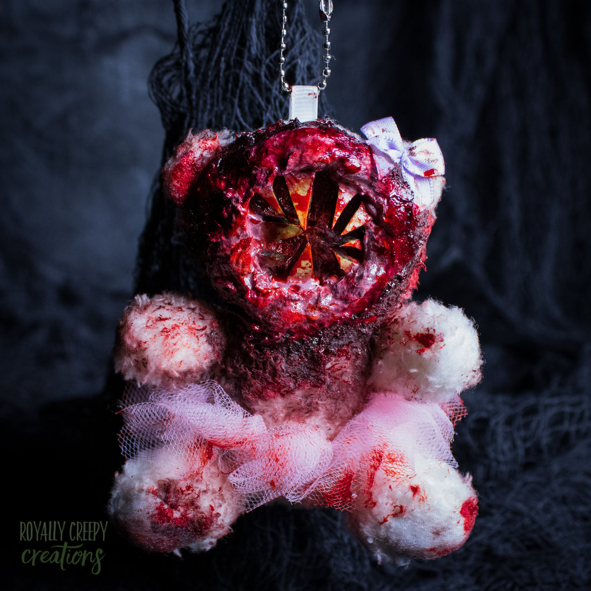 A horror-themed plush bear keychain with a bloody, gory mouth with sharp teeth taking over the whole face, fuzzy body, and wearing a pink tutu and a bow.