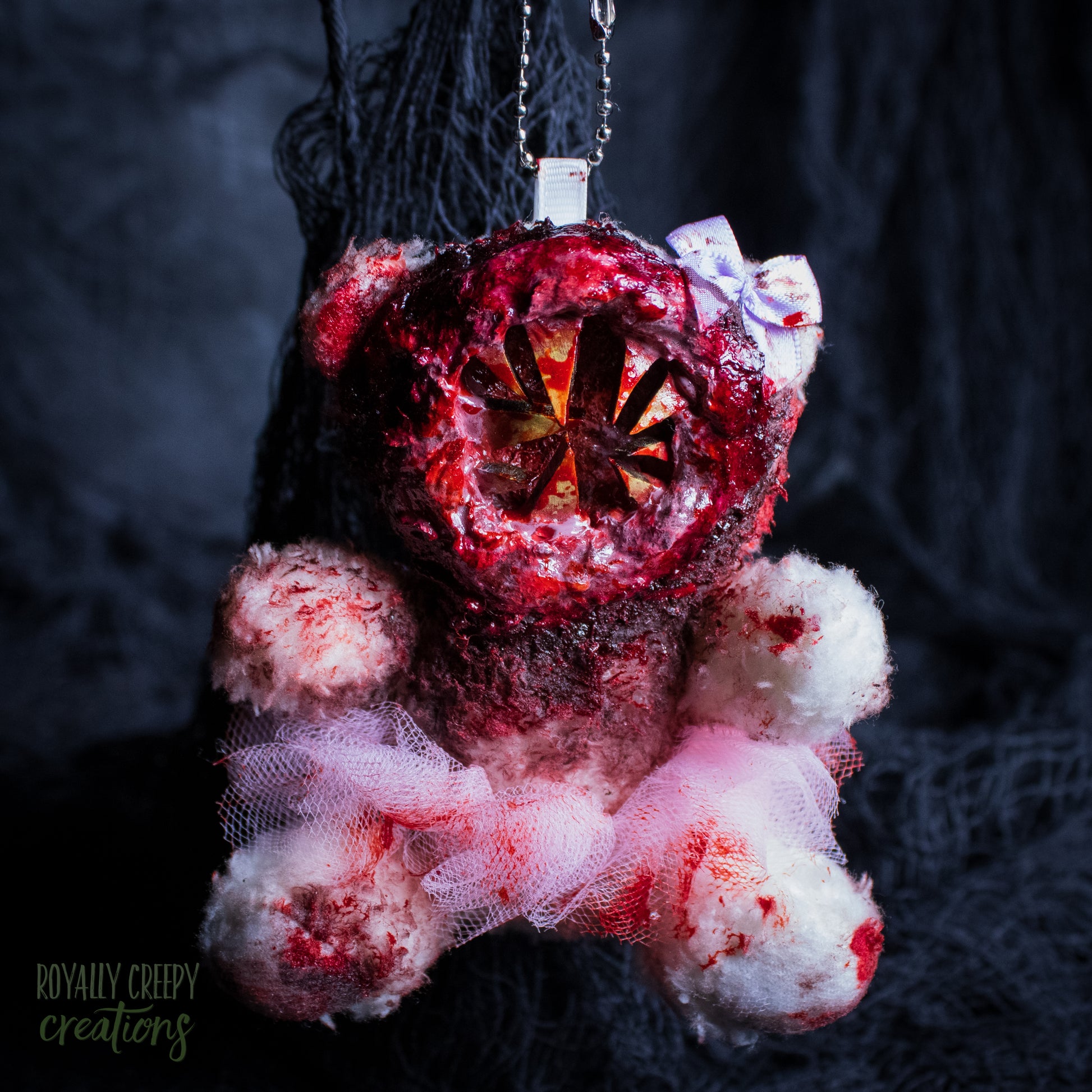 A horror-themed plush bear keychain with a bloody, gory mouth with sharp teeth taking over the whole face, fuzzy body, and wearing a pink tutu and a bow.