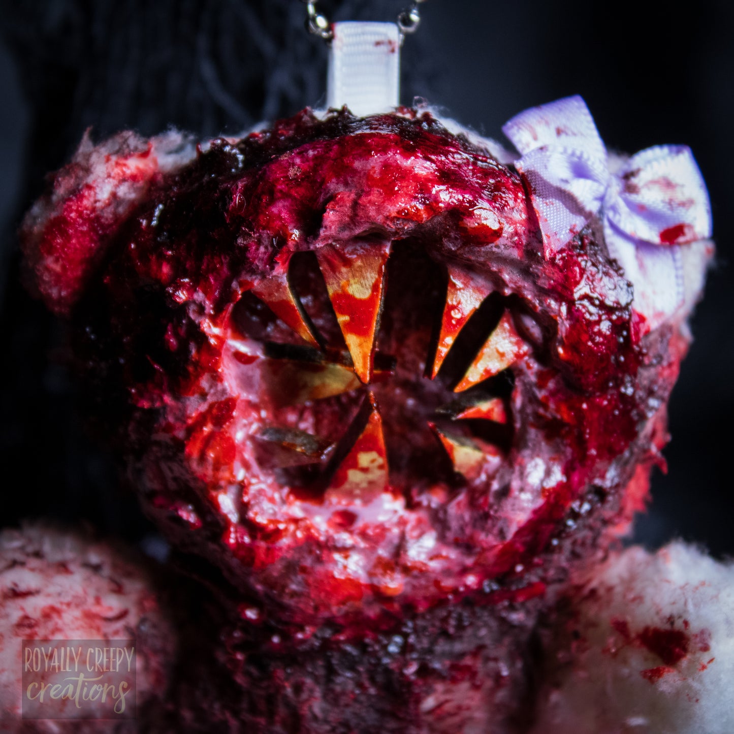 A close-up of a horror-themed plush bear keychain with a bloody, gory mouth with sharp teeth taking over the whole face, fuzzy body, and wearing a bow.