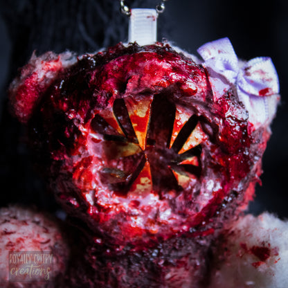 A close-up of a horror-themed plush bear keychain with a bloody, gory mouth with sharp teeth taking over the whole face, fuzzy body, and wearing a bow.