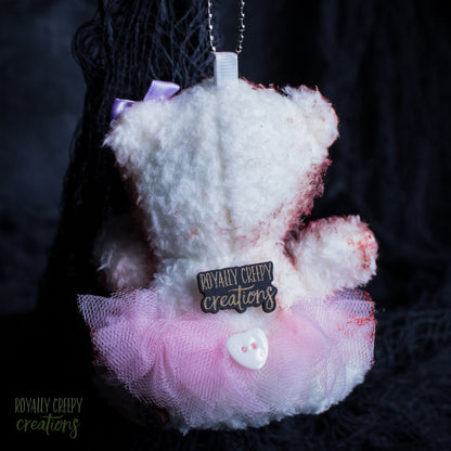 A plush teddy bear ornament white faux fur with pink tulle skirt and a small heart-shaped button, hanging against a dark black background. A brand tag reads 'Royally Creepy Creations'.