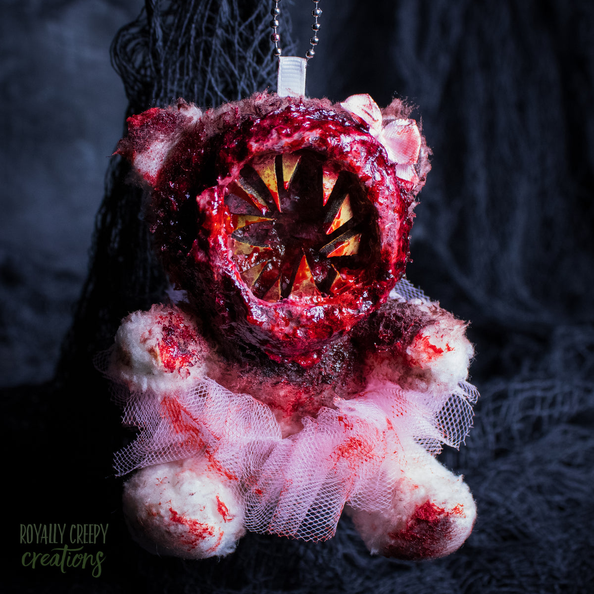A horror-themed plush bear keychain with a bloody, gory mouth with sharp teeth taking over the whole face, fuzzy body, and wearing a pink tutu and a bow.