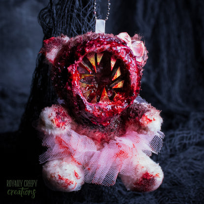 A horror-themed plush bear keychain with a bloody, gory mouth with sharp teeth taking over the whole face, fuzzy body, and wearing a pink tutu and a bow.