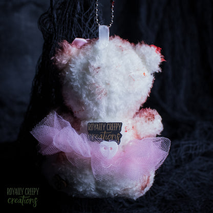 The back of a teddy bear ornament with white fluffy fur, pink tulle skirt, and fake blood splatters, featuring a 'Royally Creepy Creations' brand tag.