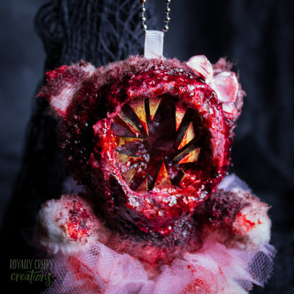 A close-up of a horror-themed plush bear keychain with a bloody, gory mouth with sharp teeth taking over the whole face, fuzzy body, and wearing a pink tutu and a bow.