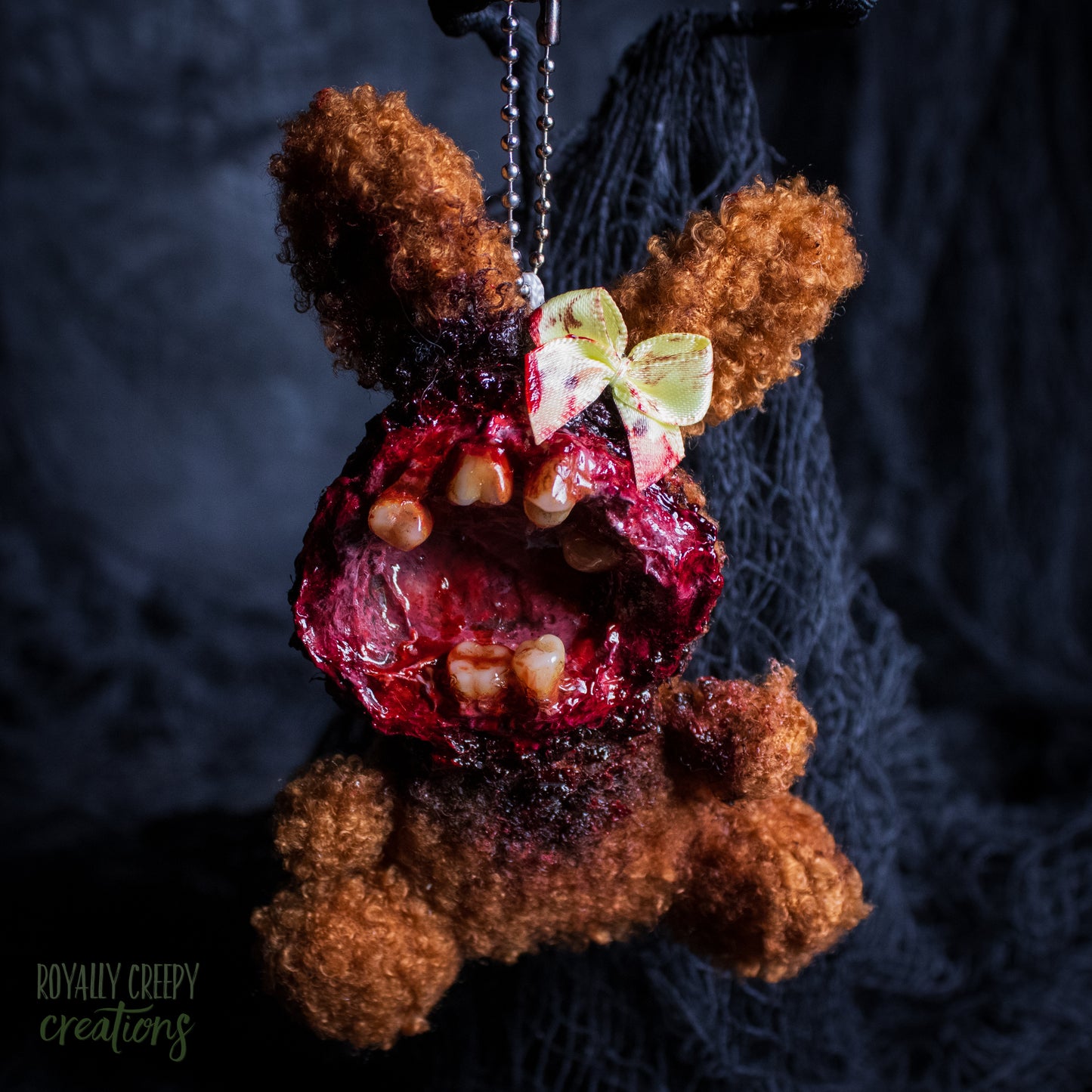 A horror-themed plush bunny keychain with a bloody, gory mouth with human teeth taking over the whole face, fuzzy body, and wearing a bow.