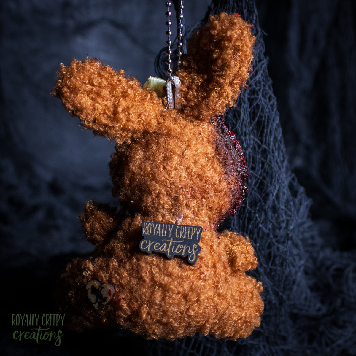 A brown curly-fiber plush bunny keychain or bag charm with a metal ball chain and a small 'Royally Creepy Creations' tag, shown against a dark textured background with black netting.