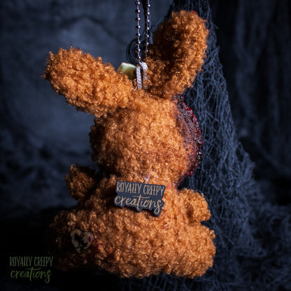 A brown curly-fiber plush bunny keychain or bag charm with a metal ball chain and a small 'Royally Creepy Creations' tag, shown against a dark textured background with black netting.