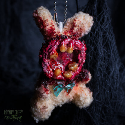 A horror-themed plush bunny keychain with a bloody, gory mouth with human teeth taking over the whole face, fuzzy body, and wearing a bow.