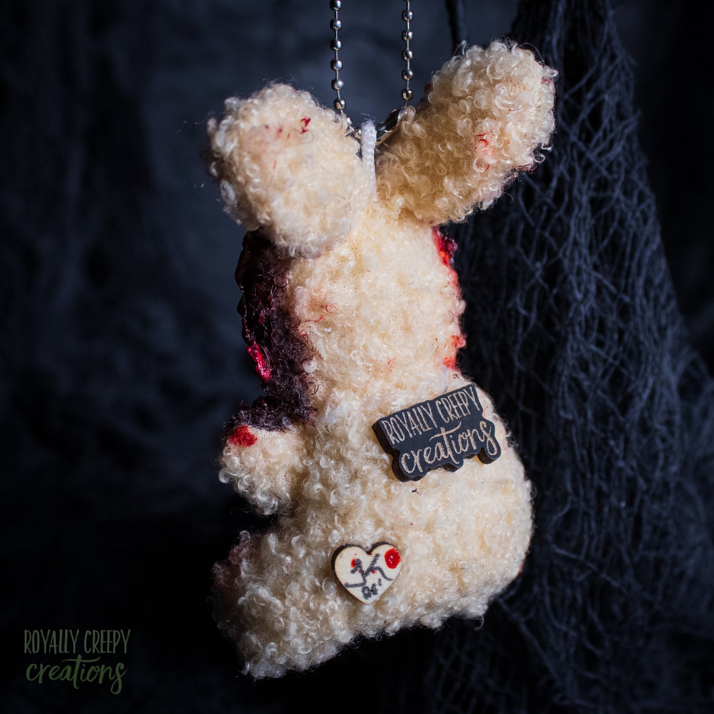 The back of a plush bunny ornament with textured cream fur, fake blood splatters, a black 'Royally Creepy Creations' patch, and a small heart charm, hanging against a dark background.