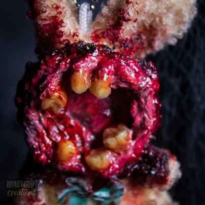 A close-up of a horror-themed plush bunny keychain with a bloody, gory mouth with human teeth taking over the whole face, fuzzy body, and wearing a bow.