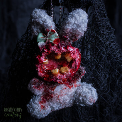 A horror-themed plush bunny keychain with a bloody, gory mouth with human teeth taking over the whole face, fuzzy body, and wearing a bow.