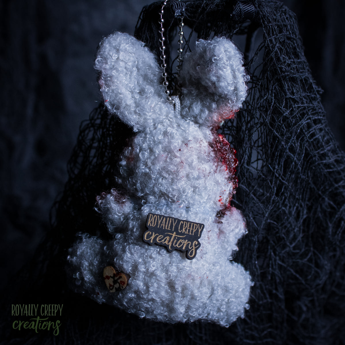 A white fluffy plush bunny keychain or bag charm with red blood-like splatter details, a metal chain, and a wooden brand tag reading 'Royally Creepy Creations,' shown against a dark netting backdrop.