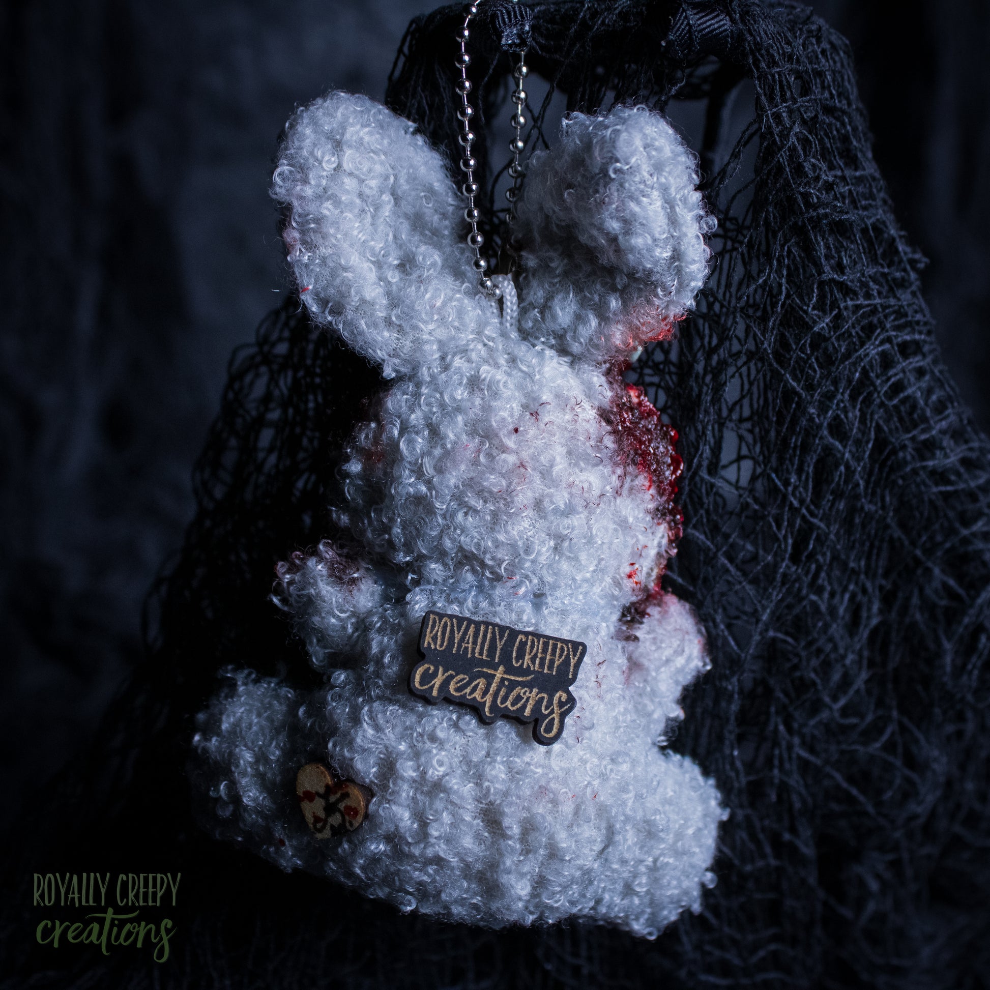 A white fluffy plush bunny keychain or bag charm with red blood-like splatter details, a metal chain, and a wooden brand tag reading 'Royally Creepy Creations,' shown against a dark netting backdrop.