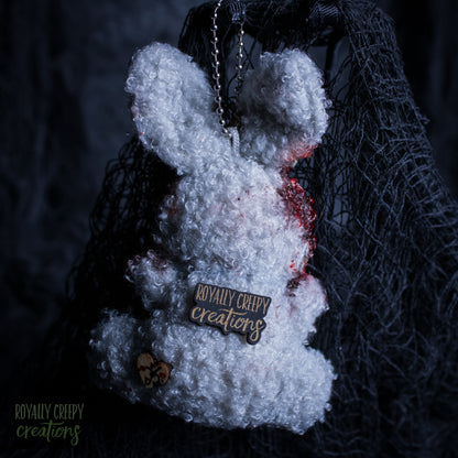 A white fluffy plush bunny keychain or bag charm with red blood-like splatter details, a metal chain, and a wooden brand tag reading 'Royally Creepy Creations,' shown against a dark netting backdrop.