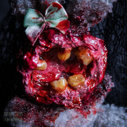 A close-up of a horror-themed plush bunny keychain with a bloody, gory mouth with human teeth taking over the whole face, fuzzy body, and wearing a bow.