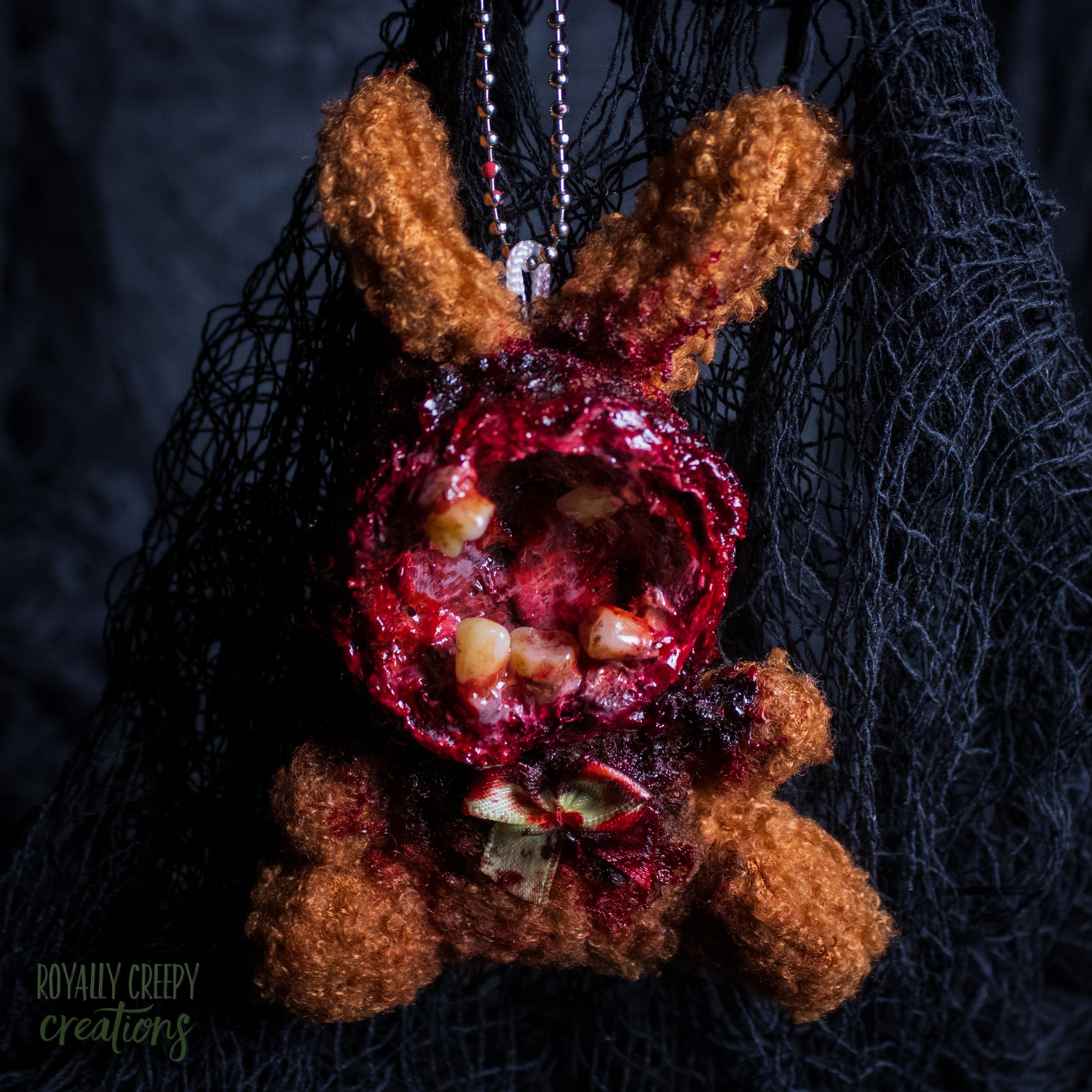 A horror-themed plush bunny keychain with a bloody, gory mouth with human teeth taking over the whole face, fuzzy body, and wearing a bow.
