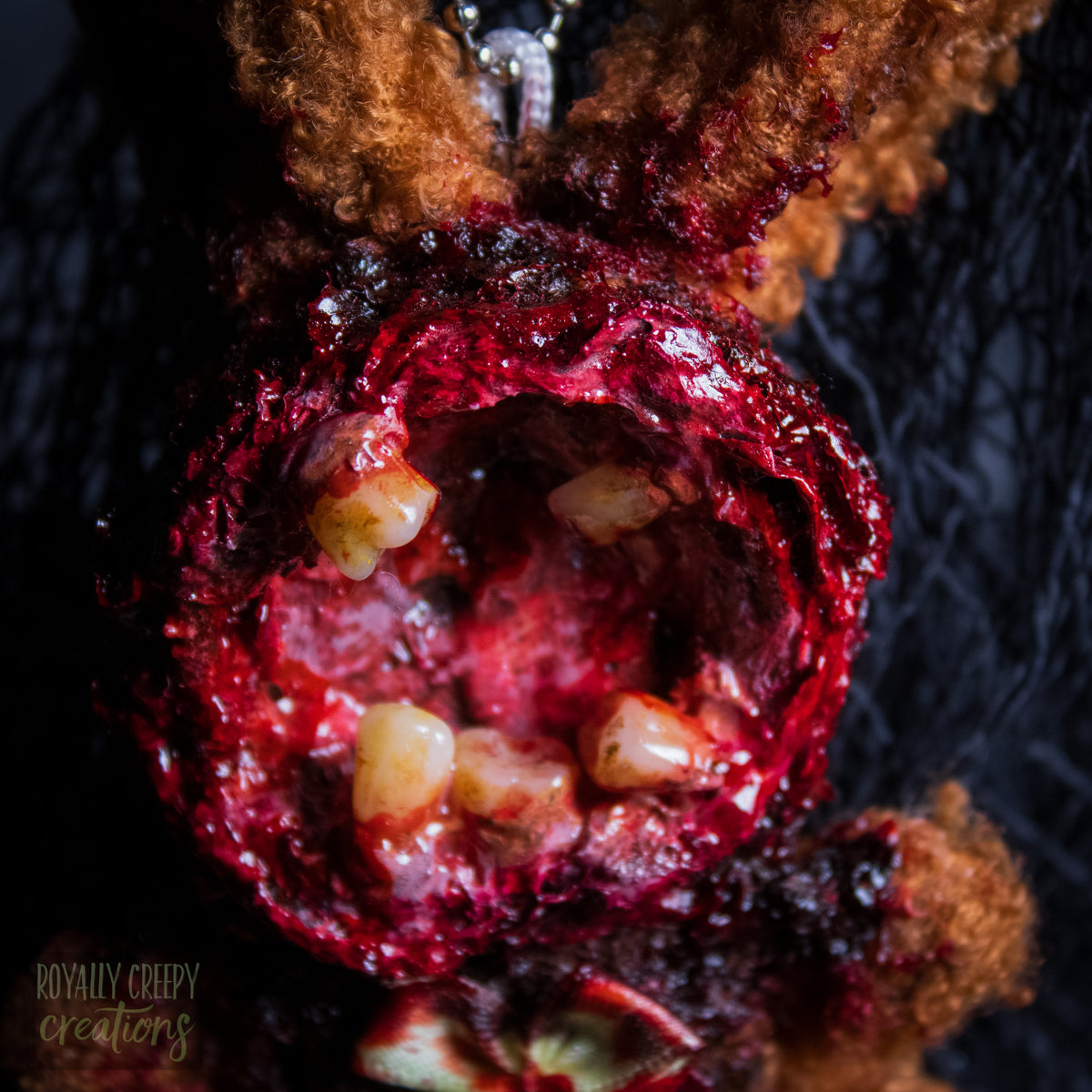 A close-up of a horror-themed plush bunny keychain with a bloody, gory mouth with human teeth taking over the whole face, fuzzy body, and wearing a bow.