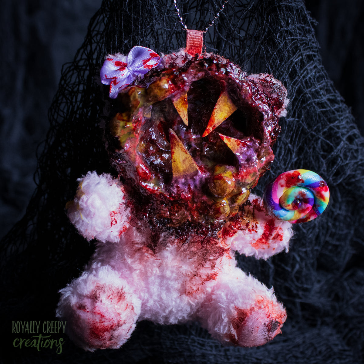 A horror-themed plush bear keychain with a bloody, gory mouth design, fuzzy body, and lollipop accessory, shown against a black netted backdrop.