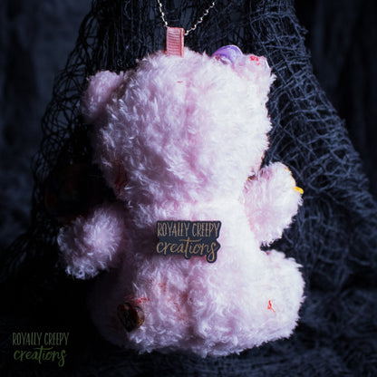 A light pink fluffy teddy bear plush keychain or ornament with a metal chain, featuring a 'Royally Creepy Creations' metal nameplate on its back, set against a dark textured background.