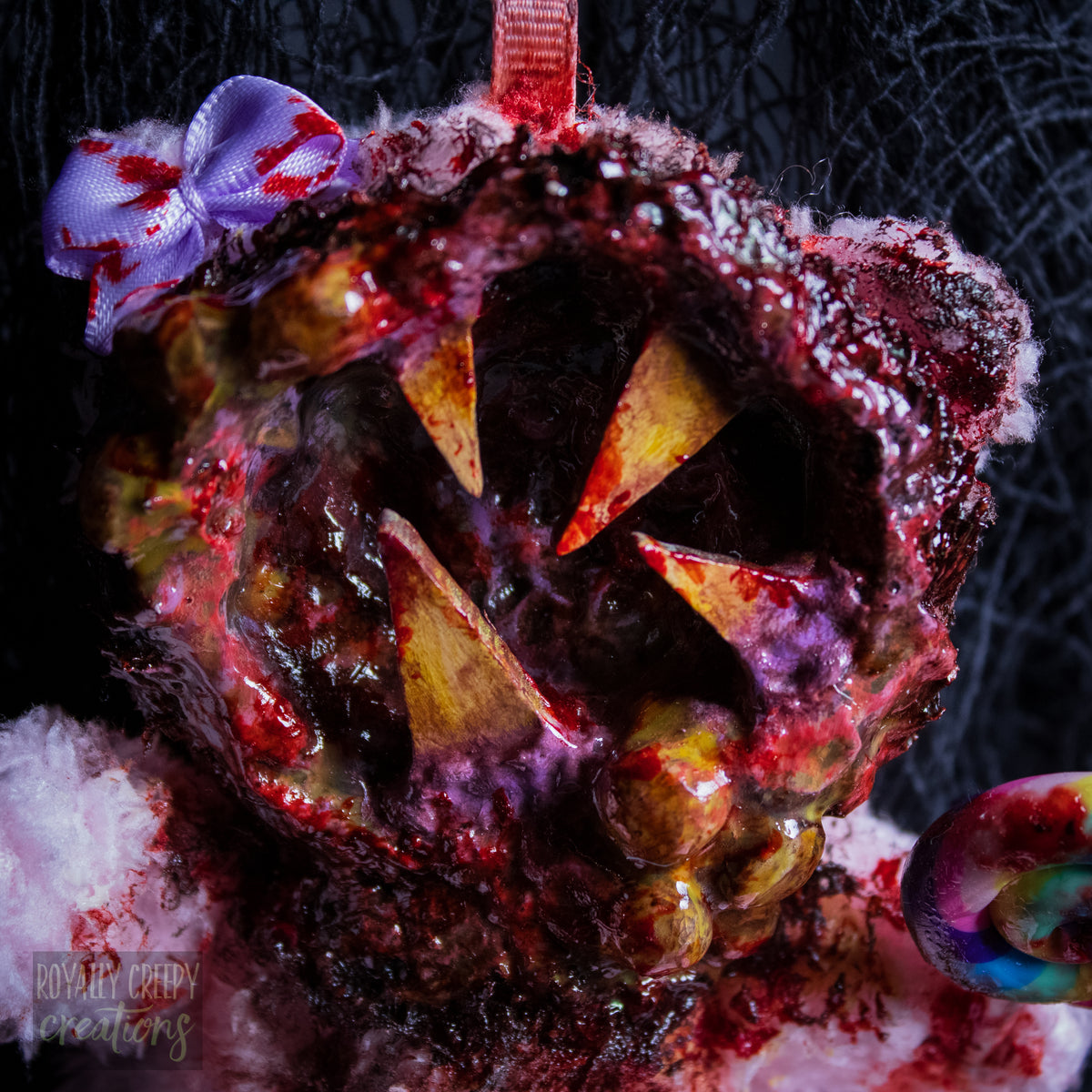 A close-up of a horror-themed plush bear keychain with a bloody, gory mouth design, fuzzy body, and lollipop accessory, shown against a black netted backdrop.