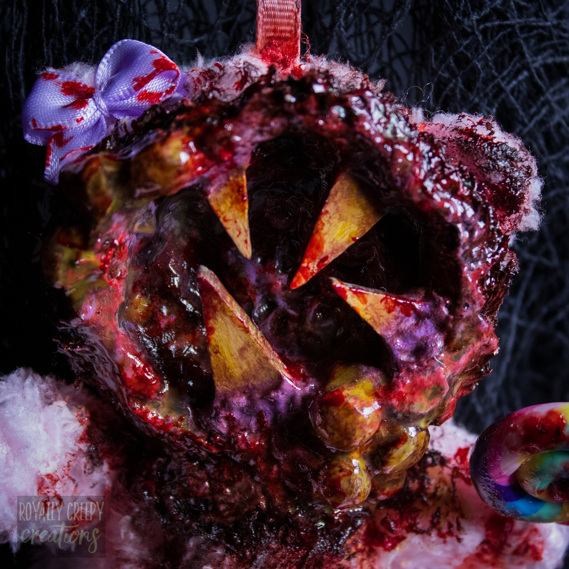 A close-up of a horror-themed plush bear keychain with a bloody, gory mouth design, fuzzy body, and lollipop accessory, shown against a black netted backdrop.