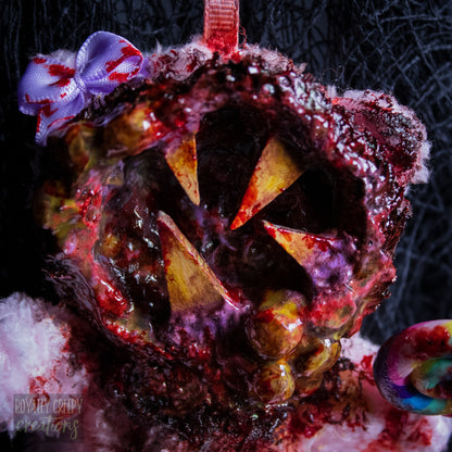 A close-up of a horror-themed plush bear keychain with a bloody, gory mouth design, fuzzy body, and lollipop accessory, shown against a black netted backdrop.