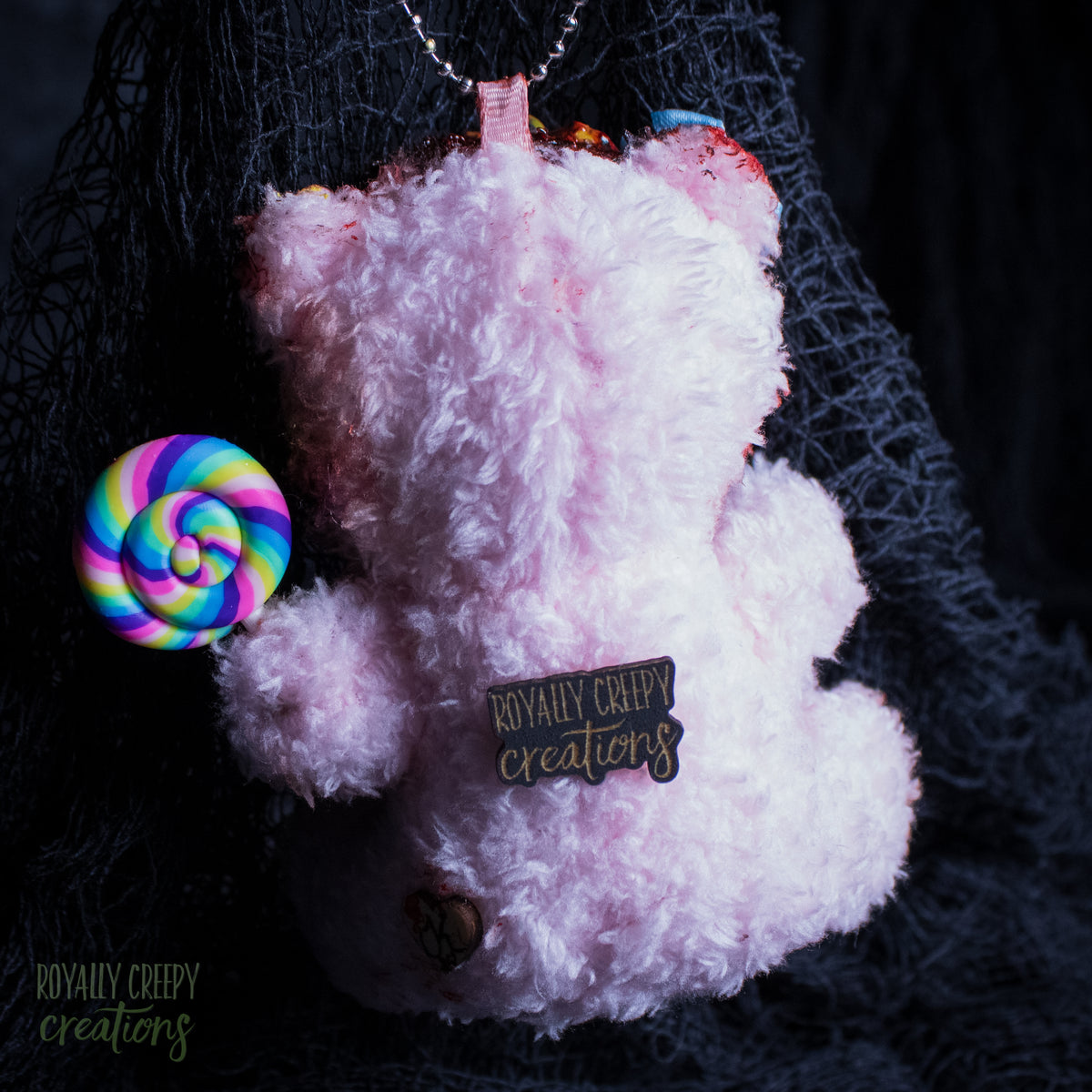 A fluffy light pink teddy bear ornament or plush accessory with a rainbow lollipop accessory and a black tag reading 'Royally creepy creations' on a dark, background with black netting.