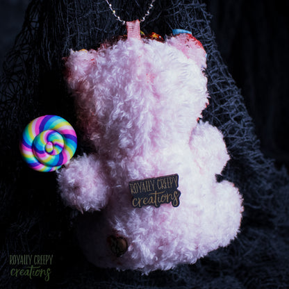 A fluffy light pink teddy bear ornament or plush accessory with a rainbow lollipop accessory and a black tag reading 'Royally creepy creations' on a dark, background with black netting.