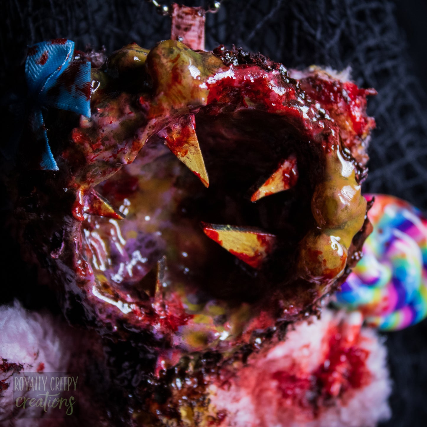 A close-up of a horror-themed plush bear keychain with a bloody, gory mouth design, fuzzy body, and lollipop accessory, shown against a black netted backdrop.