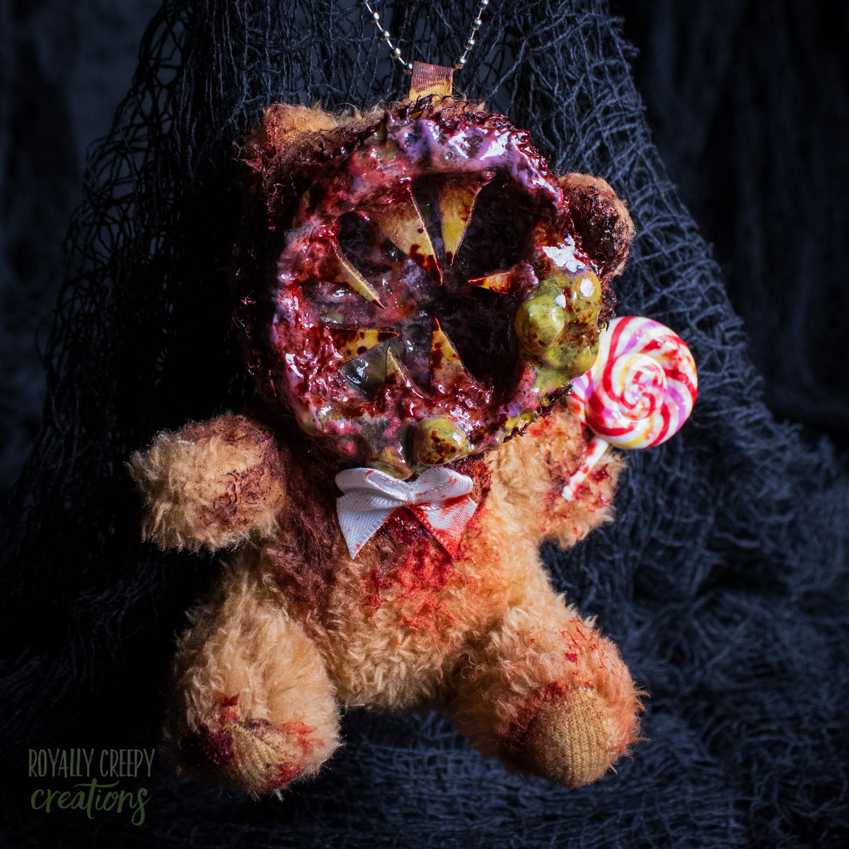 A horror-themed plush bear keychain with a bloody, gory mouth design, fuzzy body, and lollipop accessory, shown against a black netted backdrop.