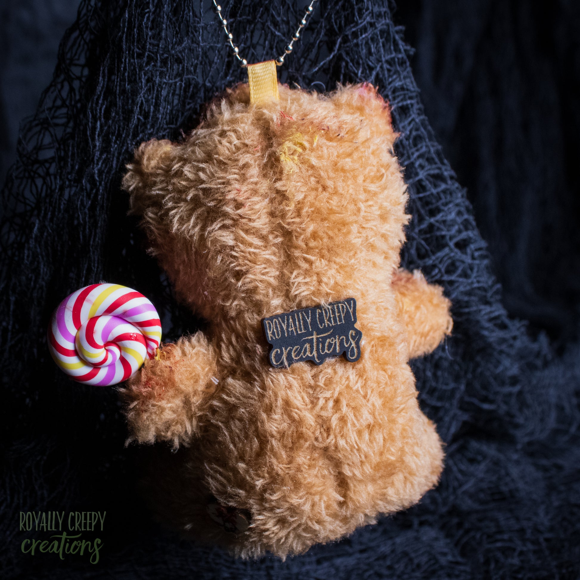 A plush teddy bear pendant necklace with a colorful striped lollipop accessory, displayed against a dark textured fabric backdrop. The bear has a black patch reading 'ROYALLY CREEPY creations' on its back and hangs from a silver chain.