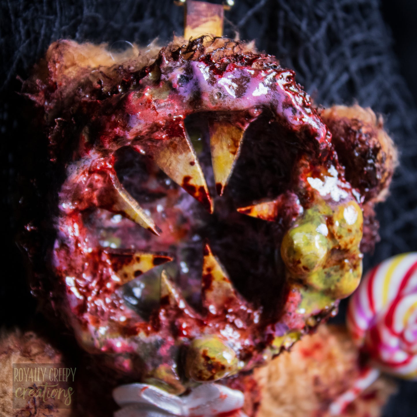 A close-up of a horror-themed plush bear keychain with a bloody, gory mouth design, fuzzy body, and lollipop accessory, shown against a black netted backdrop.