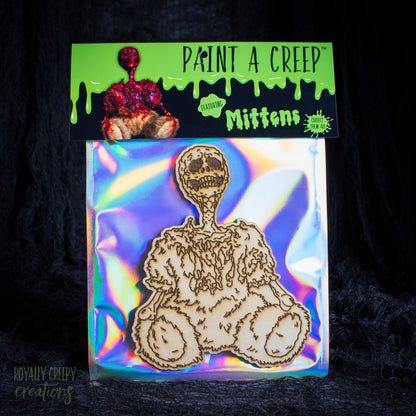 A packaged craft item showing a laser-cut wooden skeleton bearon an iridescent holographic backing card, with a branded header card reading 'Paint a Creep Mittens' against a black background.
