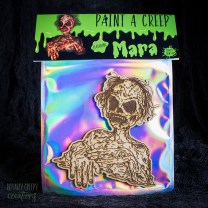 A packaged craft item showing a laser-cut wooden skeleton/creeper figure on an iridescent backing card. The top header reads 'PAINT A CREEP' and 'Mara'.
