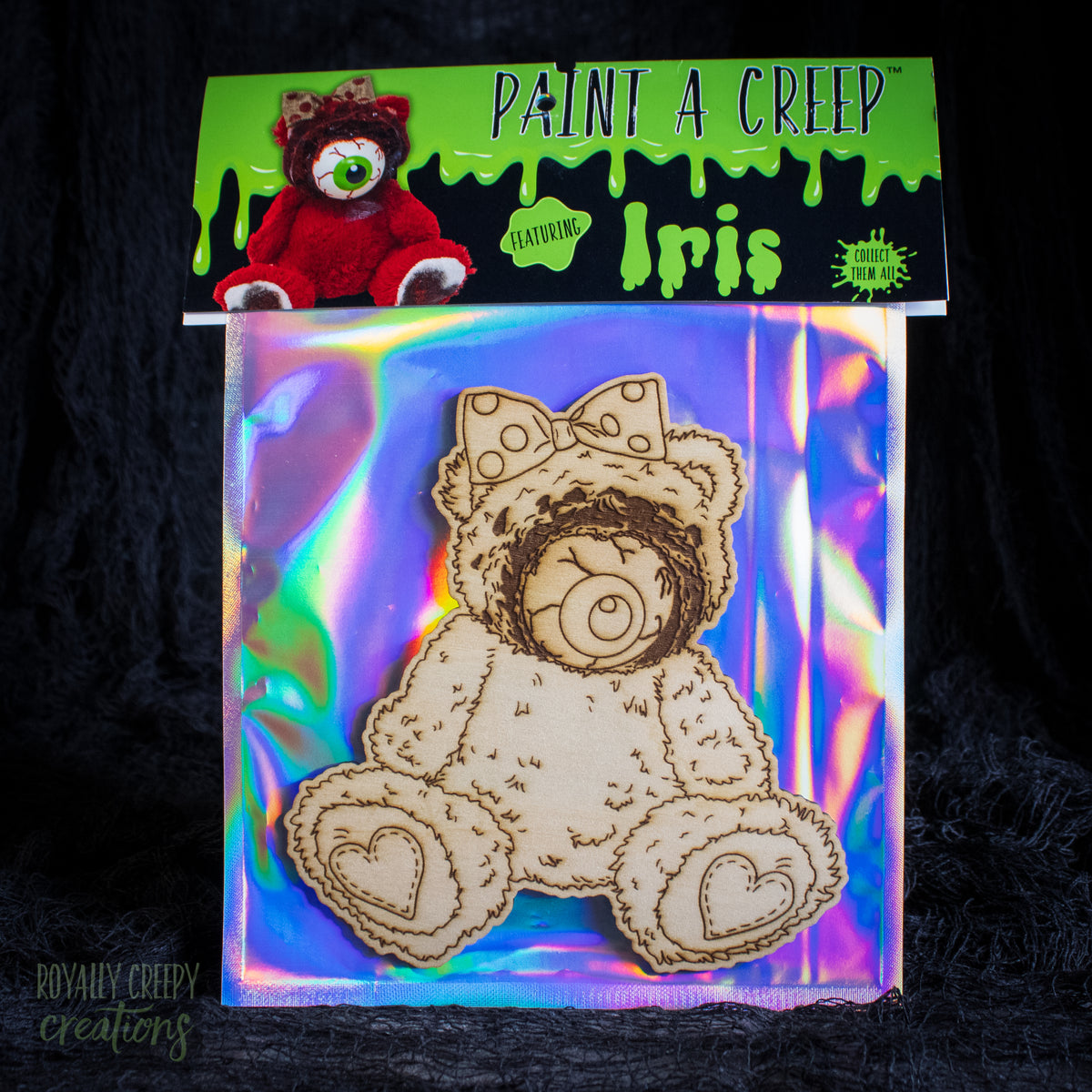 A packaged wooden bear-shaped craft piece labeled 'Paint a Creep' featuring 'Iris,' shown on an iridescent backing card with a small red plush bear reference on the header. The product is photographed against a dark textured background.