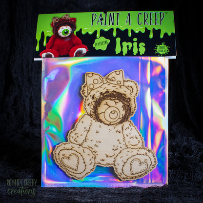A packaged wooden bear-shaped craft piece labeled 'Paint a Creep' featuring 'Iris,' shown on an iridescent backing card with a small red plush bear reference on the header. The product is photographed against a dark textured background.
